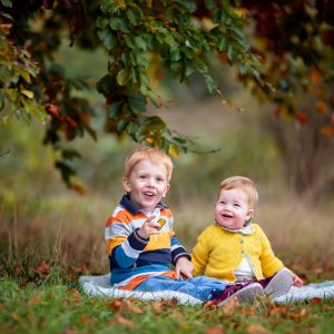 toddler and baby sitting in the autumn leaves under a tree at Wandlebury Country Park in Cambridge